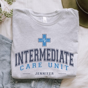 May include: A gray t-shirt with a blue and white medical cross graphic and the text "INTERMEDIATE CARE UNIT JENNIFER".