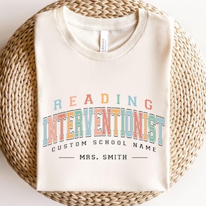 May include: Cream-colored t-shirt with the words "READING INTERVENTIONIST" in a colorful, retro font. Below, it says "CUSTOM SCHOOL NAME" and "MRS. SMITH" in a simple font. The shirt is displayed on a woven, circular surface.