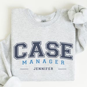May include: A light gray sweatshirt with the words "CASE MANAGER" in navy blue block letters with a white outline. Below, in smaller blue letters, is "JENNIFER". The sweatshirt is folded, with the sleeves rolled up.