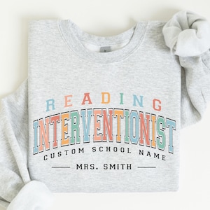 May include: A light gray sweatshirt with the words "READING INTERVENTIONIST" in colorful block letters. Below, it says "CUSTOM SCHOOL NAME" and "MRS. SMITH". The sleeves are rolled up.