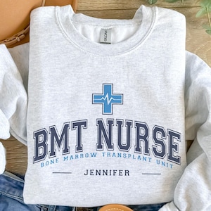 May include: Gray sweatshirt with the text "BMT NURSE" in blue and black lettering. The text "BONE MARROW TRANSPLANT UNIT" is below the main text. The name "JENNIFER" is centered below the unit text.
