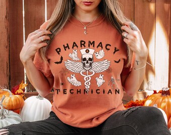 May include: A woman wearing a rust-colored t-shirt with the text "Pharmacy Technician" and a skull and snake graphic with ghosts and bats. She is wearing black jeans and black boots.