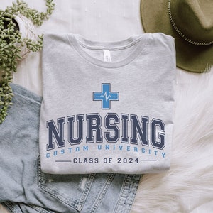 May include: Gray t-shirt with a blue medical cross and the text "Nursing Custom University - Class of 2024".