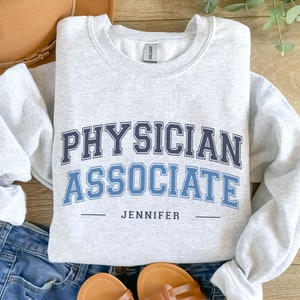 May include: Gray sweatshirt with the text "PHYSICIAN ASSOCIATE" in blue and black lettering with the name "JENNIFER" underneath.