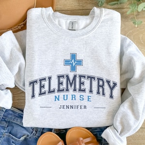 May include: A gray sweatshirt with the text "Telemetry Nurse Jennifer" in blue and black lettering. A blue cross with a heartbeat graphic is above the text.