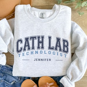 May include: Gray sweatshirt with the text "Cath Lab Technologist" in blue and black lettering. The name "Jennifer" is printed below the text.