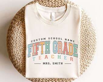 May include: A white t-shirt with the text "CUSTOM SCHOOL NAME FIFTH GRADE TEACHER - MRS. SMITH -" in rainbow colors. The text is in a retro font style.