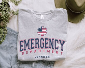 May include: A light gray t-shirt with the words "EMERGENCY DEPARTMENT" in navy blue and red lettering. The shirt features a star of life emblem with an American flag design. The name "JENNIFER" is printed below the text.