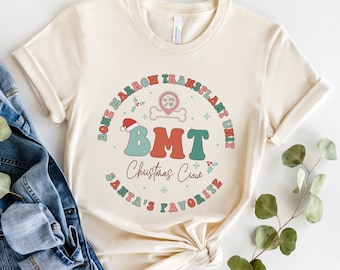 May include: A beige t-shirt with a festive design featuring the text "Bone Marrow Transplant Unit", "BMT Christmas Crew", and "Santa's Favorite". The design includes a bone marrow symbol, a Santa hat, and holly.