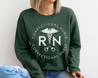 Puede incluir: Una sudadera verde oscuro con un gráfico circular blanco que dice "Correctional Nurse RN Correctional Nurse" con un caduceo y esposas en el centro.