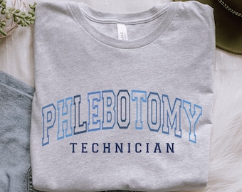 Puede incluir: Una camiseta gris jaspeada con la inscripción "PHLEBOTOMY TECHNICIAN" en azul. El texto está en una fuente de estilo universitario. La camiseta está doblada y sobre una superficie blanca.