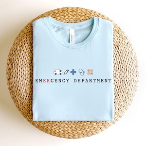 May include: A light blue t-shirt with the words "EMERGENCY DEPARTMENT" in black letters. Above the text are small illustrations of an ambulance, syringe, cross, stethoscope, and bandage. The shirt is on a woven, circular surface.