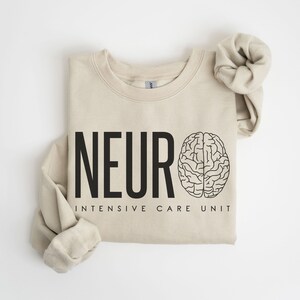 Puede incluir: Una sudadera beige con el texto "NEURO INTENSIVE CARE UNIT" y un contorno negro de un cerebro.
