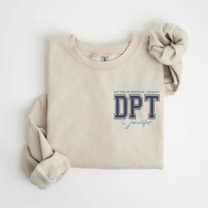 Puede incluir: Una sudadera beige con el texto "Doctor of Physical Therapy DPT Jennifer" impreso en la parte delantera.