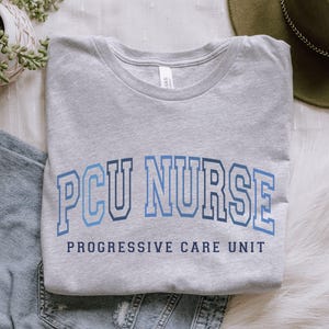 May include: A heather gray t-shirt with the text "PCU NURSE" in blue block letters. Below the text is "PROGRESSIVE CARE UNIT" in a smaller, darker blue font. The shirt is folded neatly.