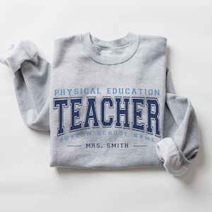 May include: Gray sweatshirt with the text "Physical Education Teacher" in blue and navy blue lettering. The text "Custom School Name" is below the word "Teacher" and the text "Mrs. Smith" is below that.