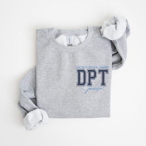 Puede incluir: Sudadera gris con el texto "DOCTOR OF PHYSICAL THERAPY DPT Jennifer" en letras azules y negras.