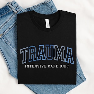 May include: Black t-shirt with the word "TRAUMA" in blue and white block letters, arched across the chest. Below the word "TRAUMA" is the text "INTENSIVE CARE UNIT" in white. The shirt is displayed on a pair of blue jeans.