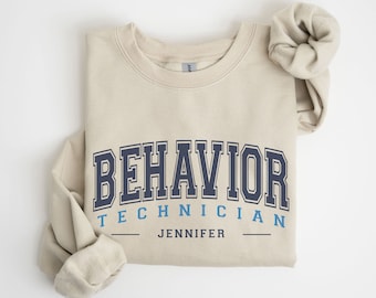 May include: Beige sweatshirt with the text "BEHAVIOR TECHNICIAN JENNIFER" in navy blue and light blue lettering.