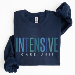 May include: Navy blue sweatshirt with the words "INTENSIVE CARE UNIT" in teal and blue lettering. The sweatshirt is folded, with the sleeves draped over the top. The Gildan brand tag is visible.