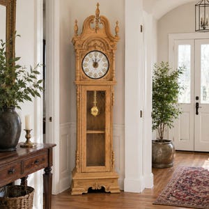 Könnte beinhalten: Eine hohe, hellfarbene Standuhr aus Holz mit dekorativer Spitze und Glastür. Das Zifferblatt ist weiß mit schwarzen römischen Ziffern. Die Uhr steht in einem Flur mit Holzboden. Die Uhr ist etwa 180 cm hoch.