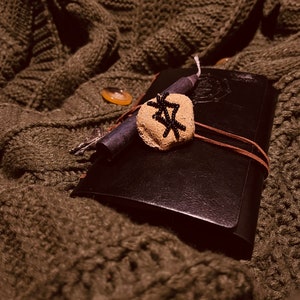 May include: A black leather journal with a leather strap, a small stone with a black symbol, and a black candle are on a green knitted sweater. The journal is closed. The stone has a rough texture.