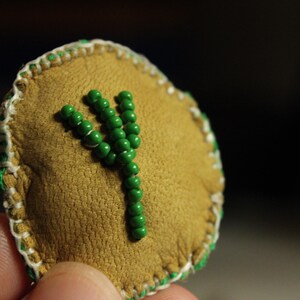 May include: A tan leather patch with a green beaded symbol. The patch has a stitched border with white and green thread. The symbol is a stylized letter or rune, made of small green beads.
