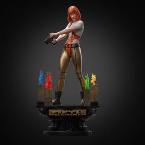 May include: A detailed figurine of a character from "The Fifth Element" movie. The figure is posed holding a gun, wearing an orange and gold outfit, and standing on a platform with the movie title. Colorful flame-like sculptures are at the base.