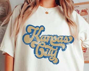 May include: A white t-shirt with a vintage-style graphic that says "Kansas City" in blue and gold lettering.