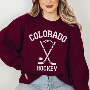 May include: A maroon sweatshirt with a white graphic design featuring the text "COLORADO HOCKEY" and a pair of crossed hockey sticks with a puck at the bottom.