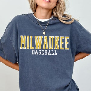 Może przedstawiać: Duża, ciemnoniebieska koszulka z żółtym nadrukiem "MILWAUKEE BASEBALL".