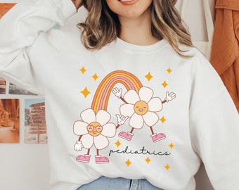May include: White sweatshirt with a rainbow and two smiling flower illustrations. The text "pediatrics" is written below the flowers.