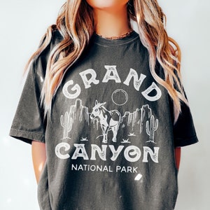 Może przedstawiać: Szary t-shirt z białym nadrukiem graficznym osła na tle pustynnego krajobrazu. Pod grafiką nadrukowany jest tekst "GRAND CANYON NATIONAL PARK".