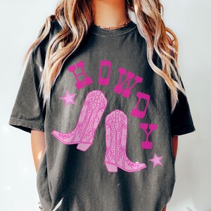 Pode incluir: Uma camiseta cinza escura com um gr&aacute;fico rosa de botas de cowboy e a palavra "Howdy" em letras rosas.