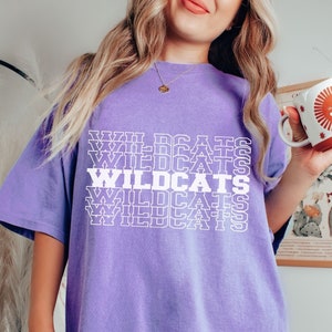 May include: A purple oversized t-shirt with the word "WILDCATS" repeated in white text. The shirt is a relaxed fit and has a crew neck.