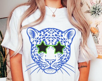 May include: White t-shirt featuring a blue and white leopard print design wearing green and black star-shaped sunglasses.