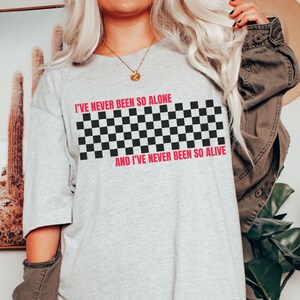 May include: A gray t-shirt with a black and white checkered pattern and the text "I've never been so alone and I've never been so alive" in pink.