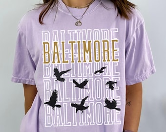 May include: A lavender t-shirt with a graphic print of the word "Baltimore" repeated four times in white with a gold outline.  There are black silhouettes of birds flying between the words.