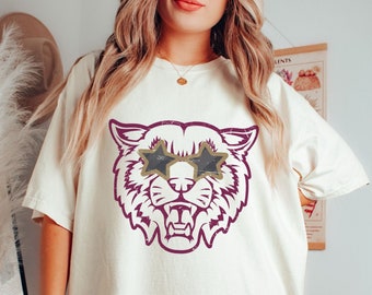 May include: A white t-shirt with a vintage-style graphic of a purple wildcat wearing star-shaped sunglasses.