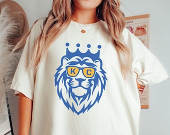 May include: A white t-shirt with a blue lion wearing yellow sunglasses and a gold crown. The sunglasses have the letters "KC" on them.