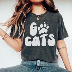 May include: A dark gray t-shirt with a white graphic that says "Go Cats" with a paw print in the "O". The model is wearing blue jeans and white sneakers.