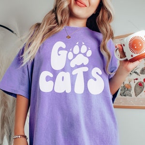 May include: A purple t-shirt with a white graphic that says "Go Cats" with a paw print in the "O".