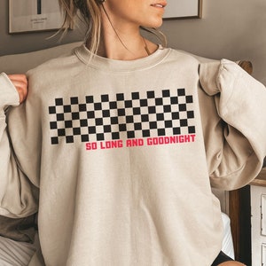 May include: Beige sweatshirt with a black and white checkered design across the chest. The text "SO LONG AND GOODNIGHT" is printed in red below the checkered pattern. The sweatshirt is a neutral color and has long sleeves.