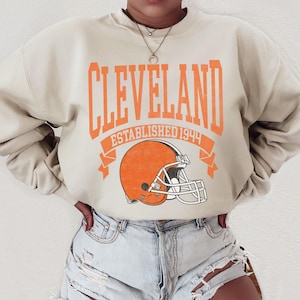 以下が含まれることがあります： オレンジ色の文字で「CLEVELAND」と書かれたベージュのスウェットシャツ。下には「ESTABLISHED 1944」と書かれたオレンジ色のバナーと、オレンジ色のフットボールヘルメットがあります。クルーネックのスタイルです。