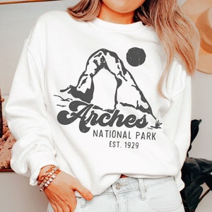 May include: White sweatshirt with a black graphic of Delicate Arch in Arches National Park, Utah. The text "Arches National Park Est. 1929" is printed below the graphic.