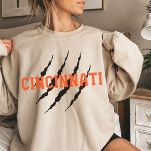 May include: A beige sweatshirt with a black and orange graphic print of the word "CINCINNATI" with claw marks.