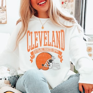 以下が含まれることがあります： オレンジ色のブロック文字で「CLEVELAND」と書かれた白いスウェットシャツ、フットボールヘルメットのグラフィック、そして「ESTABLISHED 1944」の文字。スウェットシャツはライトブルーのジーンズと合わせています。背景にはディスコボールとスマイリーフェイスのクッションがあります。