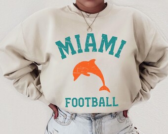 May include: A beige sweatshirt with the text "MIAMI FOOTBALL" in teal and orange. A stylised orange dolphin is jumping above the text.