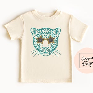 May include: A white t-shirt with a teal blue leopard print design wearing gold star-shaped sunglasses. The design has a distressed look.