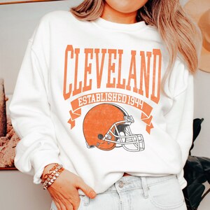 以下が含まれることがあります： オレンジ色のブロック文字で「CLEVELAND」、その下に「ESTABLISHED 1944」とフットボールのヘルメットのグラフィックがプリントされた白いスウェットシャツ。クルーネックのスタイルです。